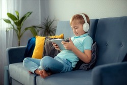 A child boy sits on a sofa and plays a game on a digital tablet and watches cartoons. Modern children's and educational technologies.
