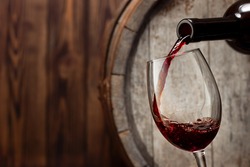 red wine pouring from bottle into glass with old wooden barrel as background