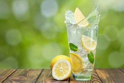 lemonade in glass with splash on wooden table and green blurred background. Summer refreshing drink. Cold detox water with lemon