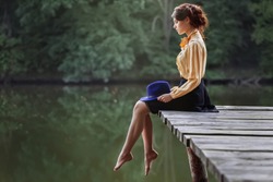 Side view of dreaming girl sitting on jetty forest and river on background. Sad girl depressed on bridge by the lake