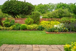 Backyard English cottage garden, colorful flowering plant and green grass lawn, brown pavement and orange brick wall, evergreen trees on background, in good care maintenance landscaping in park 