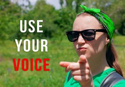 Girl shows a finger and looks at the camera with Use Your Voice text sign on green nature background. Vote elections concept. Make the political choice, your vote matters.