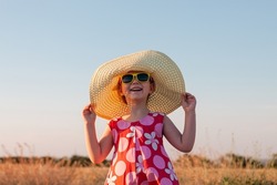 Child girl in straw hat dress in wheat spikelets field. Smiling kid in sunglasses on sunset countryside landscape. Family farming agriculture environment ecology concept. Cottagecore style aesthetic.