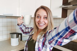 A young beautiful caucasian smiling blonde woman in the plaid shirt with a white cup of coffee or tea takes a selfie in the morning on background of the kitchen. Happy cheerful girl going to work