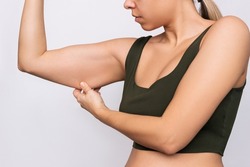 Cropped shot of a young caucasian blonde woman grabbing skin on her upper arm with excess fat isolated on a gray background. Pinching the loose and saggy muscles. Taking care of the body