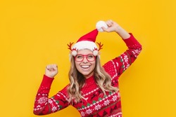 A young caucasian joyful smiling blonde girl in a Christmas red deer sweater, glasses with deer antlers and a Santa Claus hat is happy with her hands up isolated on a color yellow background