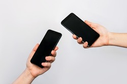 Two hands of two women holding mobile phones with blank black screens, empty copy space for design isolated on a gray background. Internet connection, File transfer, information exchange