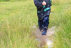 Child in a raincoat and rain boots runs through puddles. Water splashes