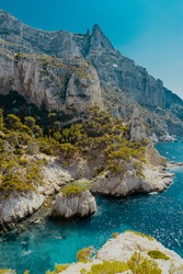 The hazy cliffs of the Calanques in France, Cote d'Azur with the deep blue sea around a small beach hidden amongst the rocks. The midday sun creates a harsh lighting condition over the trees.