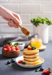 A hand pours honey on classic American pancakes in a stack with fresh berries on a blurry light gray background. The concept of a healthy, delicious and nutritious breakfast. Vertical orientation.