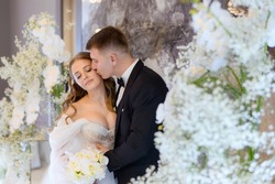 Beautiful newlyweds pose and kiss on the background of a stylish photo area decorated with white flowers and greenery