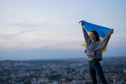 Beautiful young girl athlete conquered the top and holds the Ukrainian flag at dusk on the background of the cityscape