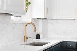 Sink detail shot in a luxury kitchen with herringbone backsplash tiles. white marble countertop, and gold faucet.