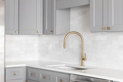 A kitchen sink detail shot with a gold faucet, marble backsplash, grey cabinets, and gold hardware.