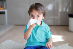 Cute toddler boy blowing nose into tissue paper at home. Mixed race Asian-German baby concept for coronavirus, Covid-19 sickness or allergy.