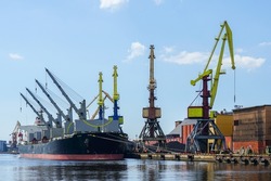 Large bulk carrier with open holds in harbor quay, shore cranes and coal piles, blue sky background