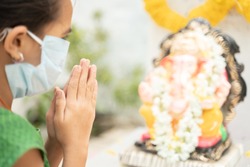 Girl Kid in medical mask praying by closing eyes in front of Lord Ganesha during Ganesha or vinayaka Chaturthi festival - concept of festival celebrations during covid-19 or coronavirus pandemic