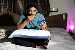 young man with headphones enjoying music videos on mobile phone by eating snacks at night- concept of unhealthy lifestyle, watching online web series and weekend relaxation.