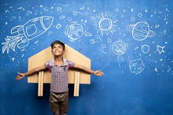 happy kid with cardboard rocket on back with space, universe and planets doodle drawing on wall - concept showing of childhood dream about astronaut or scientist