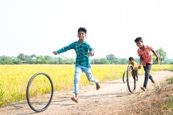 happy cheerful Indian village kids playing with tyre wheel rolling near paddy field rural street - concept of entertainment, holidays and leisure activities.