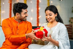 Happy young husband giving present to wife during festival celebration at home - brother giving gift to sister during raksha bandhan.