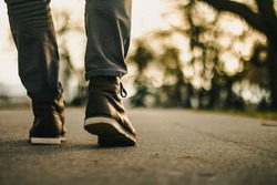 Walk in urban city. Close up view on man's legs in jeans and brown leather boots with sunlight against nature blur background.