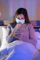 A Thai Asian woman is wearing a long-sleeved sweater and wearing a mask. She is raising her hand as if coughing and sitting on her cell phone in bed in the bedroom. Concept of Detention Corona Virus