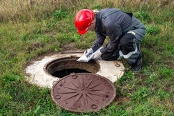 A male plumber in overalls and a helmet near a water well writes down the measurements taken and the readings of the water meter.