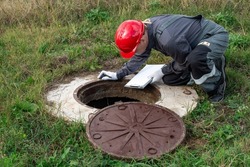A man working plumber in overalls bent over a water well fixes the measurements made and the readings of the water meter.