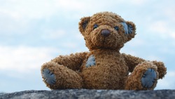 Cute little brown teddy bear plush toy sitting on stone outdoors on blue sky background.
