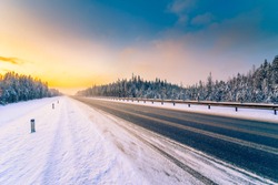 Sunrise on a clear winter morning, an empty country road to snowfall passing through a coniferous forest. View from the side of the road. Russia, Europe. Beautiful nature. Orange-blue toning effect.