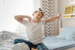Cute little kid boy wake up in the morning in his bed, stretching hand rise up to the air while sitting in sunny bedroom with big window on background.