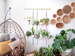 Multiple macrame plant hangers with indoor houseplants and pot planters are hanging from a metal pole. Boho basket wall decor and wicker egg chair are use to add character to the cozy bohemian room.