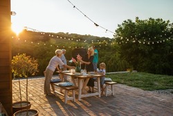 Whole family participates in the organization of dinner. Three female members setting dining table outdoors and male members barbecuing near them