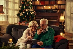 Two happy and cheerful senior sitting on the sofa using together the same tablet or technology device the christmas day - senior people having fun and sending kiss to family