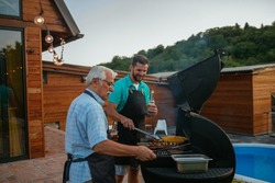 Barbecue. Leisure, food, family and holidays concept. Senior father and son wearing aprons, drinking beer and roasting meat on barbecue in the backyard