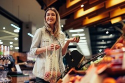 Smiling young woman standing in grocery store and holding apple, choosing freshness fruits and putting into mesh bag