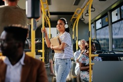 Hispanic student with earphones traveling in public transport. Smiling girl is standing at aisle while commuting by bus.