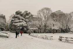A city Park with snow covered trees and benches surrounded by low buildings and people taking photos