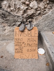 A piece of cardboard left in the street, asking people to take an image of an invisible man where there are two old shoes 