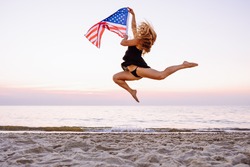 Side view of young active woman holding american flag and jumping on beach. Visa, green card, travel concept