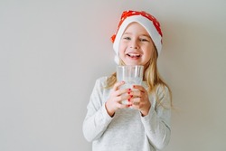 Portrait of happy laughing little girl in pajama and santa hat with glass of milk over light background