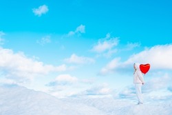 Woman holding red heart-shaped balloon behind back while standing on snow covered hill against beautiful blue sky with puffy clouds. Valentines Day Banner with copy space for text