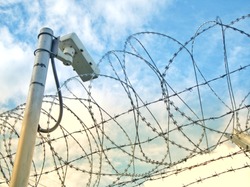 security or surveillance camera hanging behind barbed wire fence as a wall in a controlled place or prison with a blue sky background, security concept  