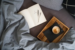 Black cup of coffee on the notebook on the wooden tray and open book with lavender flowers on the bed with grey blanket and black and white pillows. Morning ritual. Breakfast in bed.
