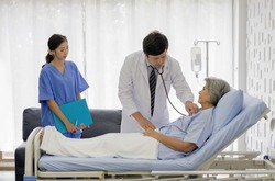 Senior female patient and doctor. Asian doctor use a stethoscope to check the heartbeat of the elderly patient. Young nurse stand next to the bed, holding document folder.