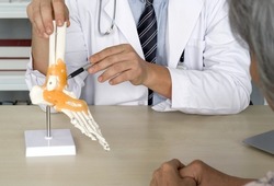 Orthopedic Surgeon in white gown and stethoscope pointing at human skeleton foot ankle bone joint anatomy model, present to the elderly patient about treatment of the ankle with surgery.