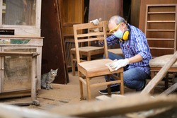 Senior Asian carpenter man is sanding wooden chair in his own garage style workshop at retirement home while his cat is watching for hobby with copy space