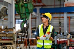 Asian engineer worker is using overhead crane hoist to carry raw materials inside metal sheet manufacturing factory for heavy industry