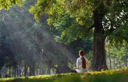 Back of woman relaxingly practicing meditation in the forest to attain happiness from inner peace wisdom serenity with beam of sun light for healthy mind wellbeing and wellness soul concept